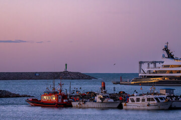 Barcos atracados en un puerto tranquilo al amanecer o atardecer, con un cielo de tonos pastel y reflejos en el agua.