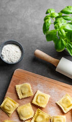 Homemade pasta ravioli with filling on wooden cutting board on dark background with flour, basil and rolling pin.
