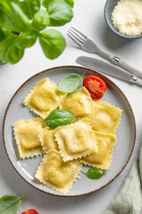 Flat lay of homemade ravioli pasta with cheese, tomatoes and basil in a plate on a light  background with  fork, herb and napkin.