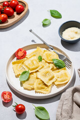Ravioli pasta with parmesan, tomatoes and basil in a plate on a light  background with shadow, napkin  and cheese.