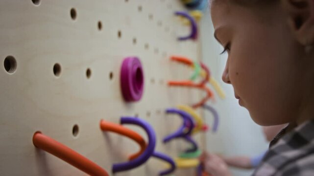 Tilt up shot of two kids focusing on threading vibrant, flexible loops onto wall-mounted pegboard in interactive play environment