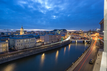 A beautiful twilight view of a river winding through Moscow, showcasing illuminated historic buildings and a lively city atmosphere, with soft blue clouds in the sky
