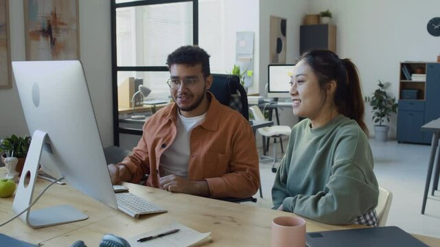 Medium slowmo shot of joyous Asian female and Arab male developers sitting together in front of computer in IT office, testing new software, giving each other high fives on successful program run