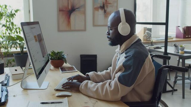 Medium side shot of black male software developer with disability, using wheelchair, sitting at desk at IT startup, typing prompts while testing new artificial intelligence image generation program