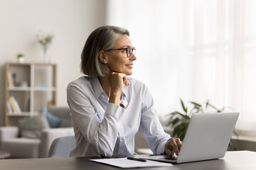 Happy dreamy mature freelancer woman in stylish eyeglasses working at home office table, sitting at laptop computer, looking away, smiling, touching chin, thinking, using pc for online communication,