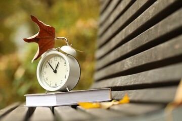 Autumn time. Alarm clock and book on bench in park, closeup