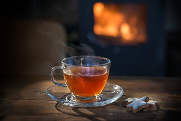 Steaming hot tea in a glass cup and cinnamon star cookies on a rustic wooden table in front of a fireplace, healthy drink in a cozy home in winter or autumn, copy space