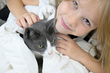 A little girl with brown hair strokes a gray cat while lying on the bed. The concept of love and care for one's neighbor.