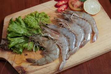 Fresh raw shrimp on wooden cutting board with lettuce, slice of red chili, tomato slice and slice lemon on the side, preparing of fresh seafood cook.