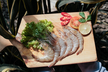 Fresh raw shrimp on wooden cutting board with lettuce, slice of red chili, tomato slice and slice lemon on the side, on background green plants, appear to be used for seafood cooking