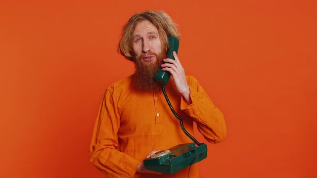 Caucasian man talking on wired landline vintage telephone of 80s, advertising proposition of conversation, online shopping, hotline service operator. Redhead young guy isolated on orange background