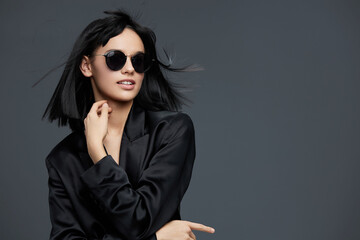 Beautiful woman in black jacket and sunglasses posing against a gray background, exuding confidence...