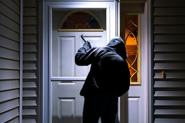 A man in a hoodie sweatshirt holds a crowbar as he tries to break into a suburban home in the evening.