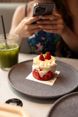 Breakfast in cafe with a piece of strawberry cake on a grey plate with iced matcha latte. Girl taking a photo of her food.