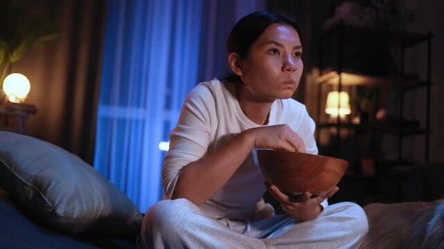 Addicted focused young asian woman looking at screen while watching interesting TV program film movie and eating popcorn at home Enchanted brunette girl cannot take her eyes off at late night indoors