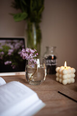 A branch of lilac in a glass of water. A white square bubble candle is lit. A book is on the wooden table.