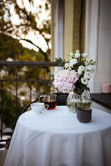 Bouquet of ranunculus blooms in a glass vase standing on a white table with cup of tea on a french balcony in the evening with street lights on its background in spring