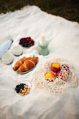 Summer picnic on a white sheet. Cup of coffee, strawberries, blueberries, nuts, peaches in a wicker bag, candle and photo camera. Morning sunrise light