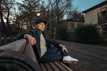 Young Man in Trendy Urban Casual Wear Relaxing on Park Bench During Autumn Evening with Vintage Buildings Background