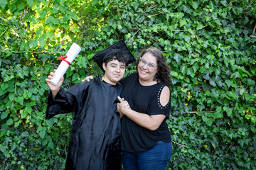 Young recently graduated boy, dressed in cap and gown, with his degree in hand, celebrating with...