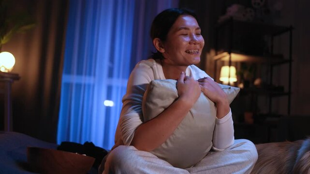 Portrait of sensitive young asian woman hugging pillow sitting on bed and watch drama film series at home indoors Beautiful sad female wipes away tears after watching love soap movie alone