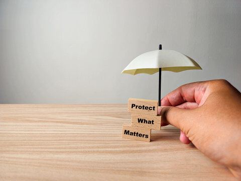 A hand holds a miniature umbrella over wooden blocks reading "Protect What Matters," symbolizing security, care, and the importance of safeguarding life&rsquo;s most valuable assets.