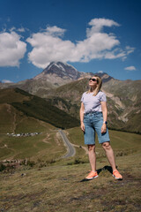Fototapeta premium A tourist girl on the background of Mount Kazbek in Georgia near the village of Stepantsminda. The girl is wearing a purple T-shirt, blue denim shorts, sunglasses. There is a mountain with a snow cap 