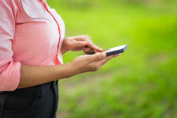 A young woman walks along a grassy path in a green forest, enjoying sunlight and nature. She relaxes, looks at her new mobile phone, and embraces the peaceful outdoor environment
