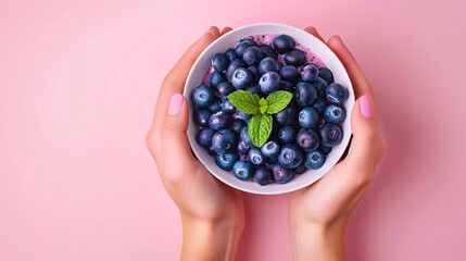A woman's hands holding a smoothie bowl with blueberries and mint on a pink background. Top view with copy space, emphasizing healthy eating.