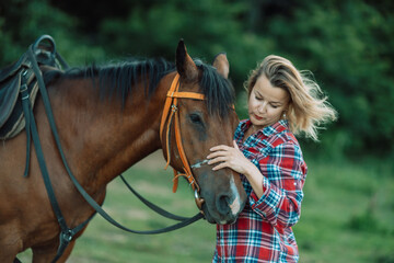 Obraz premium Happy blonde with horse in forest. Woman and a horse walking through the field during the day. Dressed in a plaid shirt and black leggings.