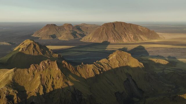 An aerial view of Thakgil, Iceland, showcasing rugged volcanic peaks and expansive shadowed plains bathed in soft golden light.