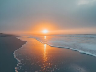 Fototapeta premium Sunrise over an empty beach with the sun reflected in the calm sea and yellow light behind a greyish-blue sky
