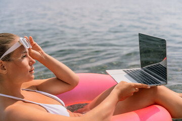 A woman is sitting in a pink float on a lake, typing on a laptop. Concept of relaxation and leisure, as the woman enjoys her time by the water while working on her laptop.