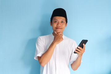 Young asian muslim man smiling holding his mobile phone while touching his chin