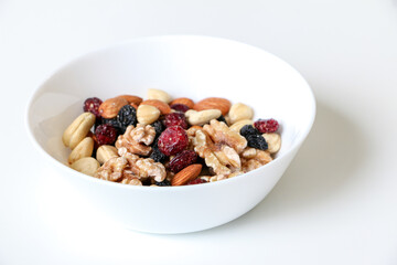 A bowl of mixed nuts and dried fruit consisting of almonds, walnuts, hazelnuts, cashew nuts, raisins and cranberries, on left side of white table with shadow