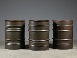 Three rusty metal barrels arranged in a row on a concrete floor with a neutral gray background, representing industrial or recycling concepts