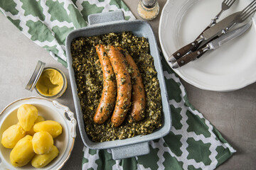 Oldenburger Grünkohl mit Pinkel Kohlwurst in Schale, Salzkartoffeln und Senf auf gedeckten grau Tisch mit grün Tuch, Teller, Besteck rustikal 