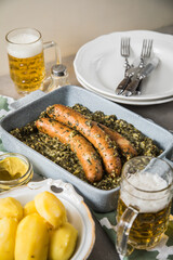 Oldenburger Grünkohl mit Pinkel Kohlwurst in Schale, Salzkartoffeln, Senf und Bier auf gedeckten grau Tisch mit grün Tuch, Teller, Besteck rustikal 