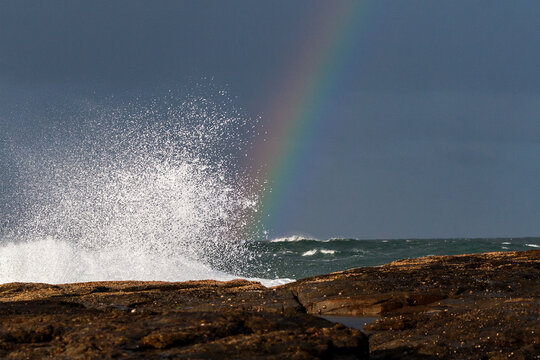 Rainbow behind crashing wave and coastal rock shelf.