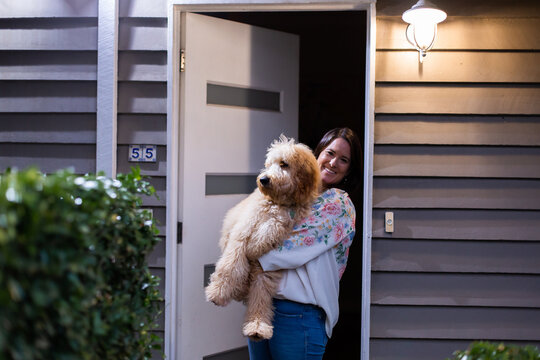 A woman smiling at the camera carrying a big brown dog at the house's front door