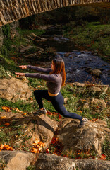 Obraz premium Young woman in tight sportswear performing a lunge exercise on rocks next to a mountain river near a stone bridge in an autumn landscape.