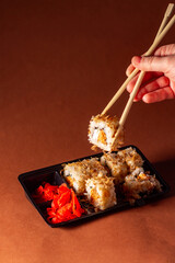 Hand with chopsticks taking portion of sushi roll. Sushi rolls with salmon cream cheese and cucumber in take away package. Food delivery concept.