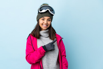 Mixed race skier girl with snowboarding glasses over isolated blue background pointing to the side to present a product.