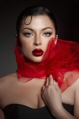 confident woman poses with striking makeup and bold red lips, dressed in a black outfit, while a flowing red scarf enhances her elegant appearance against a dark background © angel_nt