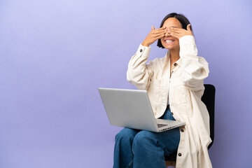 Young mixed race woman sitting on a chair with laptop isolated on purple background covering eyes...