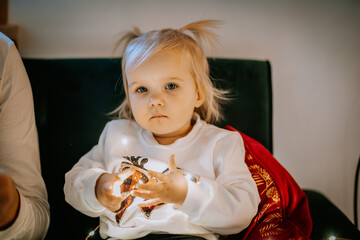 A thoughtful toddler with blonde hair sits on a chair, holding glowing fairy lights, wearing a festive outfit, and sitting near a red pillow..