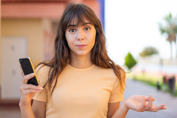Young woman using mobile phone at outdoors making doubts gesture while lifting the shoulders