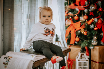 A toddler sitting on a wooden bench near a Christmas tree, wearing festive clothes with a reindeer...