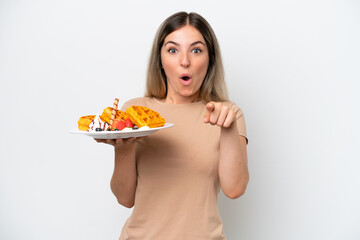 Young Rumanian woman holding waffles isolated on white background surprised and pointing front