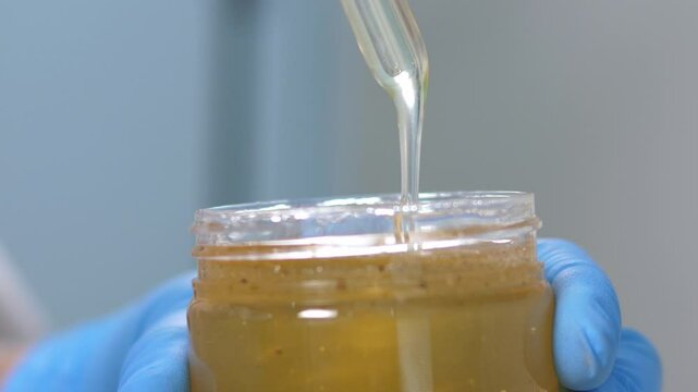 Close-up of a female laboratory assistant holding a jar of honey for analysis and quality control. A researcher wearing gloves takes a sample of honey for analysis for the presence of antibiotics.